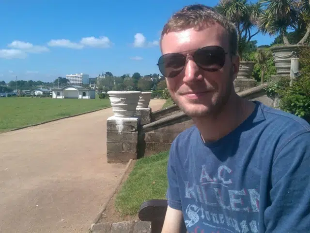 A young white man sat down outside. He is wearing a light blue t-shirt and black sunglasses.