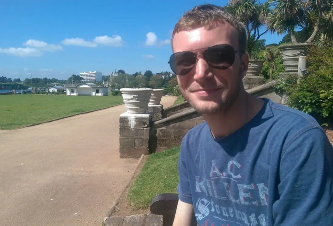 A young white man sat down outside. He is wearing a light blue t-shirt and black sunglasses.