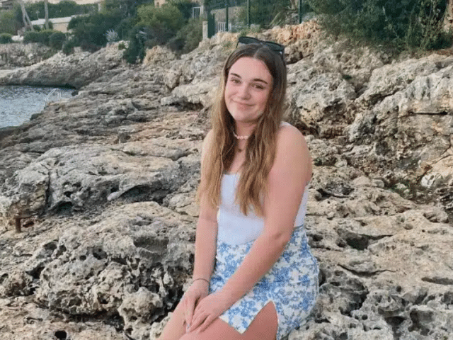 A young white women sat on rocks by a beach.