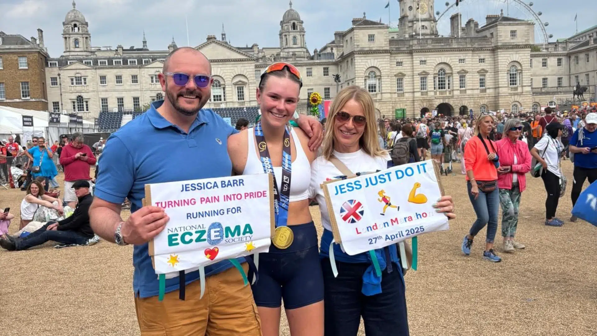 A group of three people. The woman in the middle is wearing running clothing with a London Marathon finishers medal around her neck. The people either side are holding supportive posters for the runner in the middle.